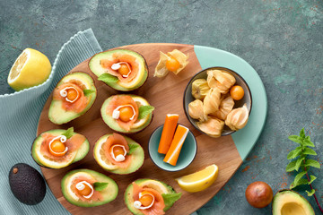 Avocado boats with smoked salmon, crab sticks, lemon, physalis and mint leaf, top view