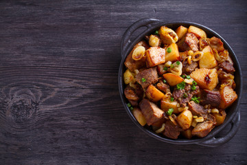 Fried beef and  potatoes with onions and garlic served in black dish on wooden background. Copy space, room for text