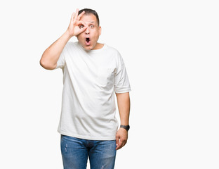 Middle age arab man wearig white t-shirt over isolated background doing ok gesture shocked with surprised face, eye looking through fingers. Unbelieving expression.