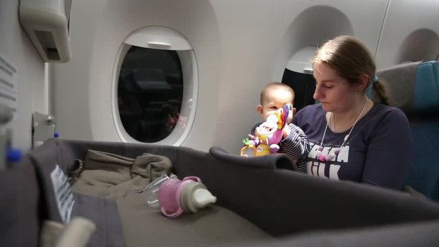 Small baby girl in the plane on mother hands play with toy. Infant child first time in flight and travel