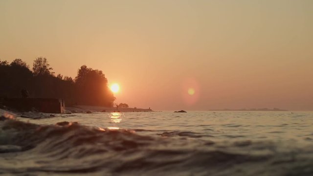 Handheld shot close up of calm river waves at the sunset under orange sky at summertime. Evening sunset and tides of river on stone shores trees and island on background.