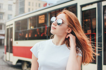 Attractive redhead smiling girl in sunglasses in casual clothes on tram background at street in city