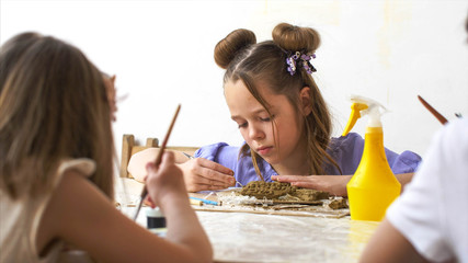 Child girl is sculpting toy from wet clay. She is using special tools on art lesson in workshop at...