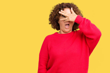 Beautiful middle ager senior woman red winter sweater over isolated background peeking in shock covering face and eyes with hand, looking through fingers with embarrassed expression.