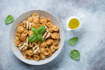 Orecchiette pasta with slices of grilled cheese and fresh green basil, flatlay on a light-grey stone background