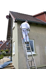 Rear view of a painter standing on a ladder and stroking the outside wall of a house with a paint roller.  