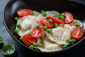 Closeup of a black bowl with ravioli, cherry tomatoes and green peas, selective focus