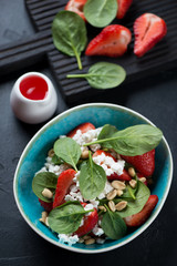 Bowl of salad with spinach leaves, strawberries, cottage cheese and nuts, vertical shot over black stone background