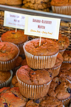 This is a color photo of homemade banana-blueberry gluten free and dairy free muffins. They have a descriptive sign on the muffins.