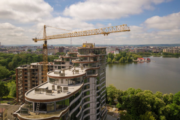 Obraz premium Apartment or office tall building unfinished under construction among green tree tops. Tower cranes on bright blue sky copy space background.