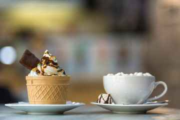 Ice cream dessert in wafer cup with chocolate cookies and and mug of coffee with marshmallows on porcelain plate on blurred colorful copy space background.