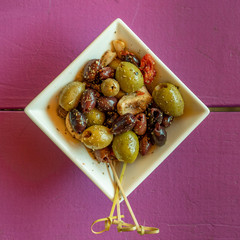 Black, red and green olives in a white bowl with garlic and toothpicks. Top view on purple table.