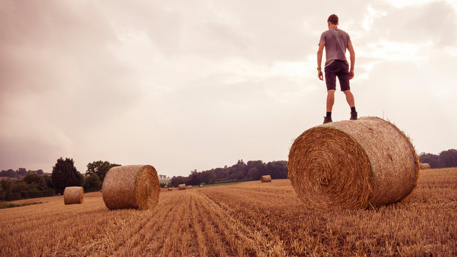 Fields, Ditzingen, Germany: Young man with backpack stands on bale of straw and overlooks the landscape while a thunderstorm is tearing in.