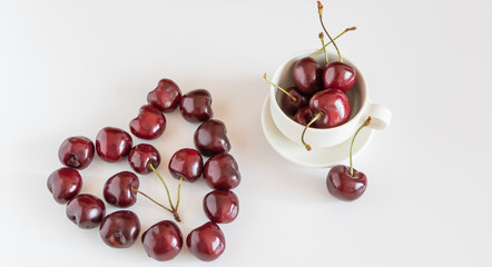 Figure of heart made of red sweet  cherries near a cup with a sweet cherries on a white background.  Top view. Copy space.