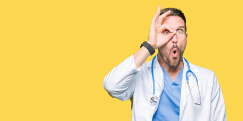 Handsome doctor man wearing medical uniform over isolated background doing ok gesture shocked with surprised face, eye looking through fingers. Unbelieving expression.