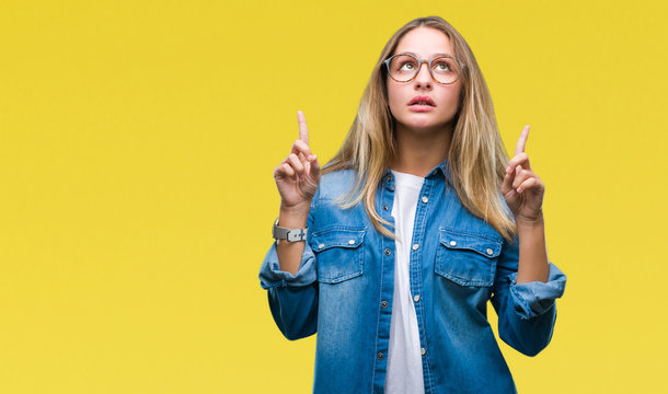 Young beautiful blonde woman wearing glasses over isolated background amazed and surprised looking up and pointing with fingers and raised arms.