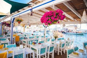 Fototapeta na wymiar View of restaurant or cafe and bougainvillea flowers on beach in Gumusluk, Bodrum city of Turkey. Aegean seaside style colorful chairs, tables and flowers in Bodrum town near beautiful Aegean Sea.