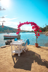 View of restaurant or cafe and bougainvillea flowers on beach in Gumusluk, Bodrum city of Turkey. Aegean seaside style colorful chairs, tables and flowers in Bodrum town near beautiful Aegean Sea.
