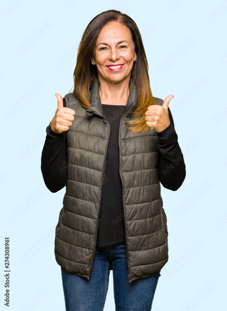 Wall mural Beautiful middle age woman wearing winter vest success sign doing positive gesture with hand, thumbs up smiling and happy. Looking at the camera with cheerful expression, winner gesture.