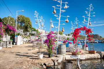 View of restaurant or cafe and bougainvillea flowers on beach in Gumusluk, Bodrum city of Turkey. Aegean seaside style colorful chairs, tables and flowers in Bodrum town near beautiful Aegean Sea.