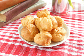 homemade small cakes profiterole choux pastry with custard next to a white cup with tea and books on a table with a rustic tablecloth in a red-white cage
