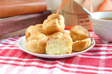Bitten homemade small cakes profiterole choux pastry with custard next to a white cup with tea and books on a table with a rustic tablecloth in a red-white cage