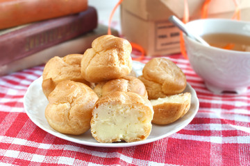 Bitten homemade small cakes profiterole choux pastry with custard next to a white cup with tea and books on a table with a rustic tablecloth in a red-white cage