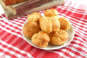 homemade small cakes profiterole choux pastry with custard next to a white cup with tea and books on a table with a rustic tablecloth in a red-white cage