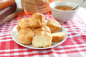 homemade small cakes profiterole choux pastry with custard next to a white cup with tea and books on a table with a rustic tablecloth in a red-white cage