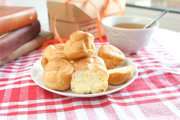 homemade small cakes profiterole choux pastry with custard next to a white cup with tea and books on a table with a rustic tablecloth in a red-white cage