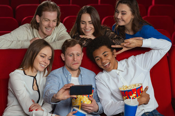 Funny company watching comedy in cinema and taking selfie on phone. Young girls and boys looking at camera, laughing, posing and making faces in movie house. Concept of free time and happiness.