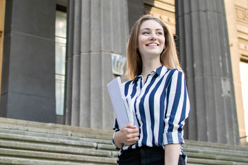 pretty student standing near the university, holding papers in her hands and smiling against the background of the college, she goes to school