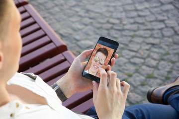 Young man sitting on bench and using her cellphone. Publishing selfie in social media site.