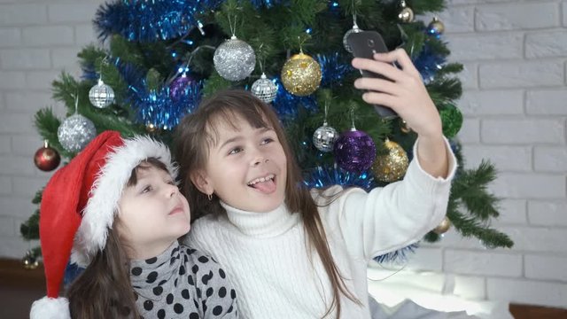 Two small girls play with faces. Cute little sisters are photographed on a smartphone against the background of a Christmas tree.