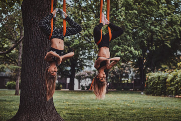 Two happy smiling girls are doing exercises upside down on the slings in the summer park.