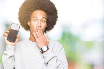 Young african american man with afro hair showing smartphone screen cover mouth with hand shocked with shame for mistake, expression of fear, scared in silence, secret concept