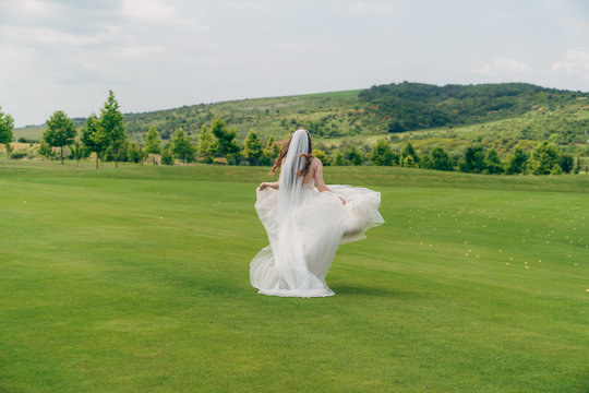 Backside view, bride runs in white wedding dress with veil on green field