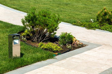 walking path of marble tiles and stone edges in the garden with a green lawn and flowerbed with plants and pine trees with a lighting device metal ground lamp.