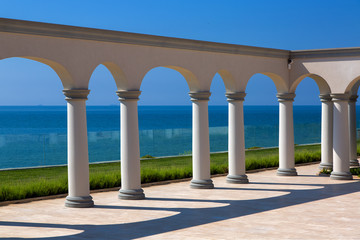 an arch with columns of white stone on sunny day and a marble floor with a lawn and a view of the sea on the coast of a luxurious mansion.
