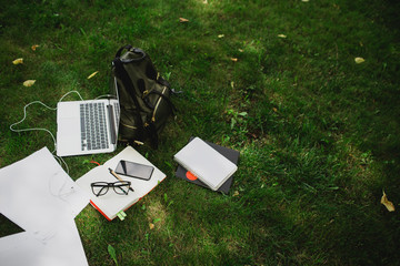 Student or freelancer tools on green grass: laptop, backpack, notebook, smartphone, empty sheets of paper. 