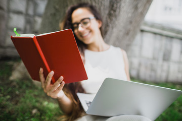 Beautiful girl student learning her lessons using her laptop and notes, sitting on green lawn in a yard of university.