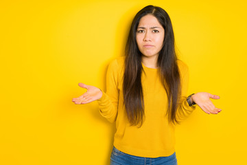 Beautiful brunette woman over yellow isolated background clueless and confused expression with arms...
