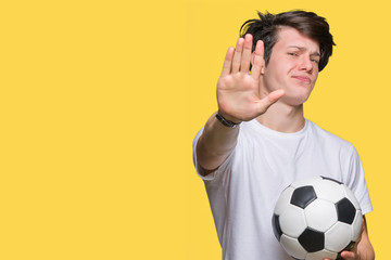 Young man holding soccer football ball over isolated background with open hand doing stop sign with serious and confident expression, defense gesture