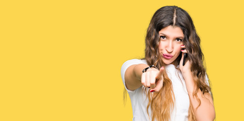 Young adult woman taking on the phone pointing with finger to the camera and to you, hand sign, positive and confident gesture from the front