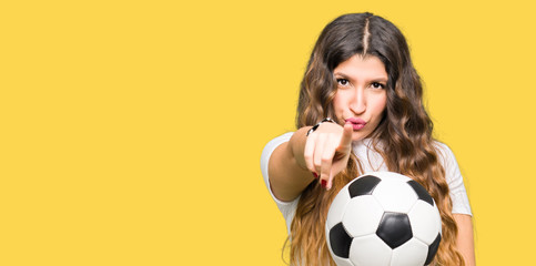 Young adult woman holding soccer football ball pointing with finger to the camera and to you, hand...