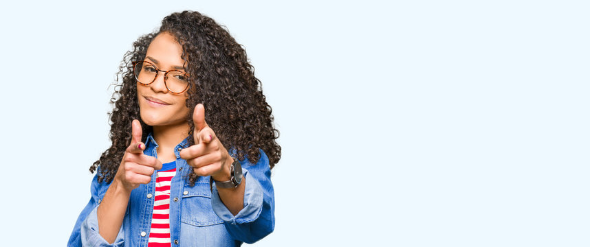 Young beautiful woman with curly hair wearing glasses pointing fingers to camera with happy and funny face. Good energy and vibes.