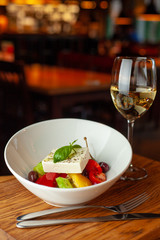Traditional fresh greek salad with cherry tomatoes, bell pepper, cucumber and feta cheese dressed with olive oil served with white wine on wooden table with blurred background