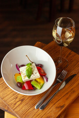 Traditional fresh greek salad with cherry tomatoes, bell pepper, cucumber and feta cheese dressed with olive oil served with white wine on wooden table with blurred background