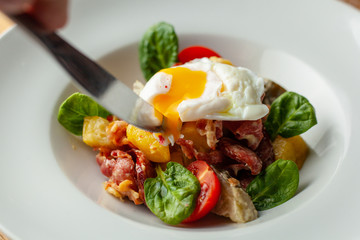 Cutting poached egg with yellow yolk on rustic salad with, tomatoes, chorizo sausage, spinach, potato and pickled cucumber served on wooden table