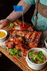 Waiter is holding a big assortment of hot meat snack for beer served on wooden dish board: big pork knuckle, BBQ pork ribs, chicken thigh kebab with mustard and sauced